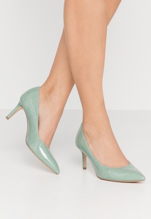 Jambes portant des escarpins en cuir verni crocodile vert menthe avec des talons aiguilles de hauteur moyenne sur fond blanc.