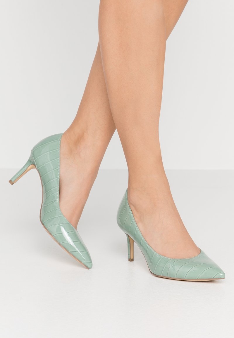 Jambes portant des escarpins en cuir verni crocodile vert menthe avec des talons aiguilles de hauteur moyenne sur fond blanc.