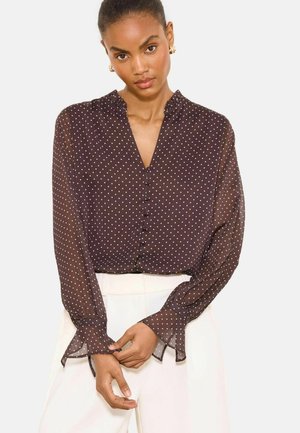 Kobieta nosząca prześwitującą ciemną bluzkę z długim rękawem w białe kropki i z guzikami, w połączeniu z białymi spodniami z wysokim stanem.