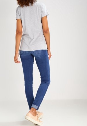 Kvinde, der står med ryggen til, iført en grå kortærmet trøje, blå opsmøgede jeans og beige sneakers på en ensfarvet baggrund.