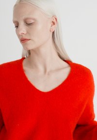 Pull rouge à col en V en tissu doux et texturé. Présente des épaules tombantes et une coupe décontractée, soulignant un design contemporain.