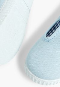 Zapatillas slip-on de lona azul claro con una suela de goma texturizada y detalle de forro a cuadros en azul. Sin marcas ni adornos visibles.