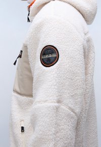 Beige fleecejacka med en rund svart och orange Napapijri-patch på överarmen och en beiget dragkedjorficka på sidan.