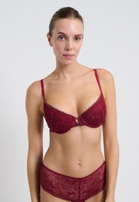 Mujer vestida con un conjunto de encaje burdeos a juego, sujetador y bragas, de pie frente a un fondo blanco liso, mirando hacia adelante.