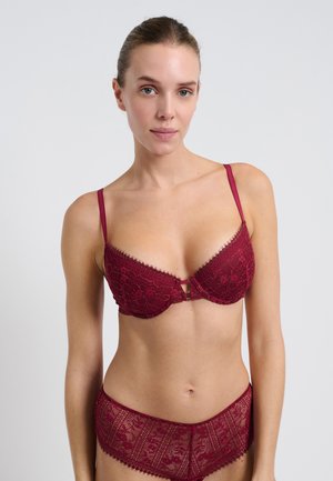 Femme portant un ensemble en dentelle bordeaux assorti, comprenant un soutien-gorge et une culotte, se tenant devant un fond blanc uni, tournée vers l'avant.