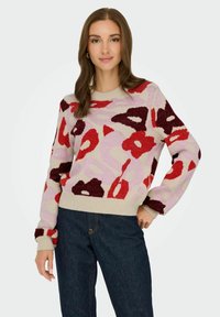Pull en tricot avec des motifs abstraits en rouge, bordeau, rose et crème. Il possède un col rond et des poignets côtelés, avec une finition texturée.