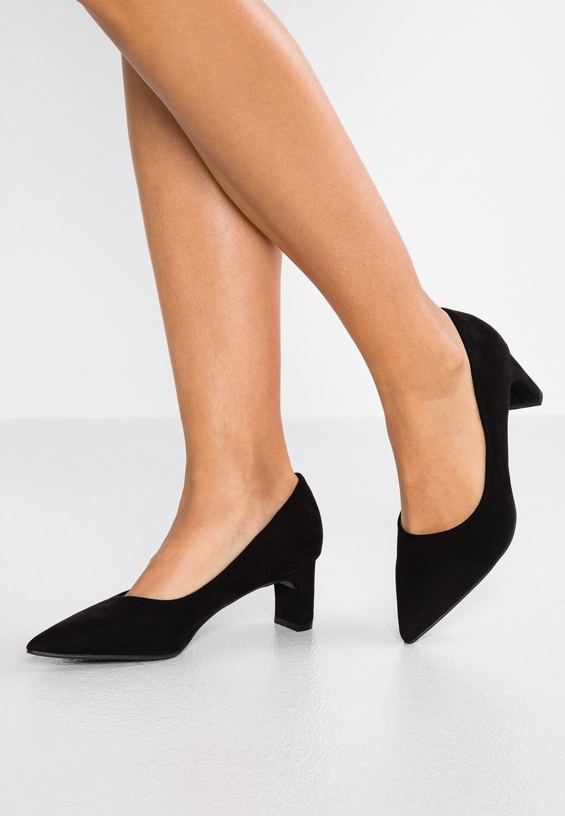 Jambes de femme portant des chaussures en daim noir à bout pointu avec un petit talon carré, sur un sol gris clair et un fond blanc.