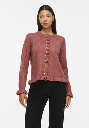 Femme portant un cardigan boutonné rose-brun avec des volants sur le devant et aux poignets, associé à un pantalon noir, debout devant un fond uni.