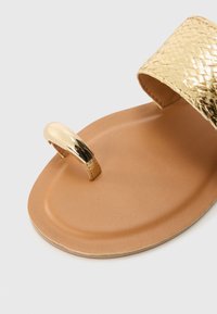 Goldfarbene, gewebte Rutsch-Sandale mit einer glatten, tanfarbenen Sohle, die eine metallische Zehen-Schleife für zusätzlichen Halt und ein strukturiertes Oberdesign aufweist.