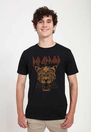 Jeune homme souriant, portant un t-shirt noir Def Leppard avec un motif tête de léopard féroce et un pantalon beige, debout sur un fond clair uni.