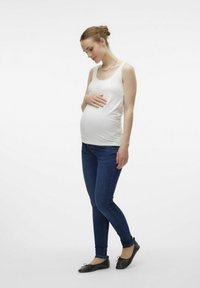 Top de maternidad en blanco, de corte ajustado; combinado con jeans skinny azules y bailarinas negras; mujer abrazando suavemente su vientre.