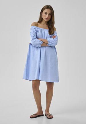 Nő áll, karjait keresztbe téve, világoskék csíkos, váll nélküli ruhát visel, fekete papucsokkal, egyszínű háttér előtt.