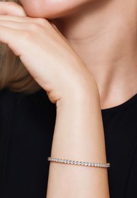 Braccialetto sottile e delicato con una fila di piccoli cristalli trasparenti incastonati in un cinturino d'argento o platino, indossato su un polso contro uno sfondo scuro.
