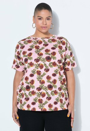 Vrouw met kort haar draagt grote ronde oorbellen en een roze shirt met een patroon van vijgen en bladeren, gecombineerd met zwarte broek met textuur.