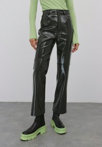 Schwarze, hoch taillierte Kunstlederhose mit glänzender Oberfläche, ausgestattet mit strukturierten Nähten und geraden Beinen, kombiniert mit klobigen grünen Plateaustiefeln.