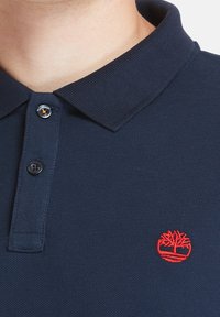 Marinblå piké skjorta med ribbad krage, med två svarta knappar och en röd broderad trädlogotyp på vänster sida av bröstet.