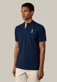 Hombre que lleva una camiseta polo azul marino con el número blanco "1" y el logo "Hackett London", combinado con pantalones beige claros, de pie frente a un fondo liso.