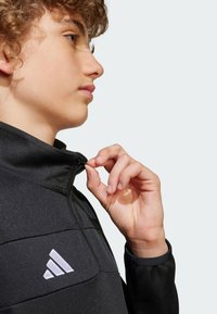 Crna jakna s patentnim zatvaračem i visokim ovratnikom, od teksturirane tkanine, s bijelim Adidas logotipom na prsima. Ruka modela prilagođava zatvarač.