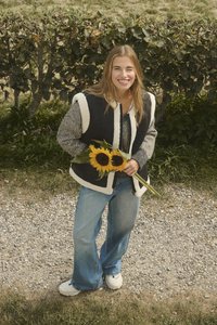 Mlada žena stoji na šljunčanom putu držeći dva suncokreta, odjevena u crno-bijeli prsluk, sivi pulover, plave traperice i bijele tenisice na otvorenom.