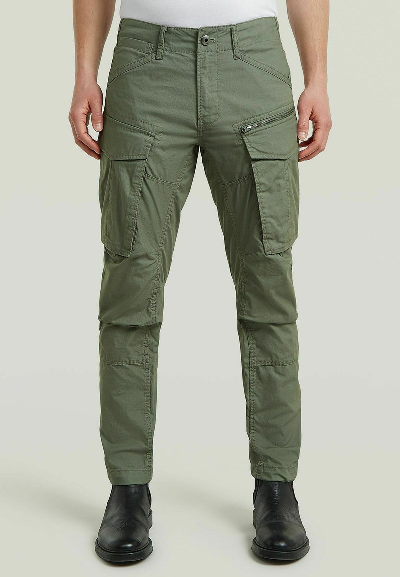 Man wearing green cargo pants with multiple pockets and black shoes, standing against a plain light background.