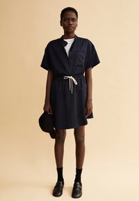 Marineblauwe jurk met korte mouwen, een geknoopte kraag, elastische tailleband en ivoorkleurige koord. Model houdt een zwarte pet vast en draagt zwarte sokken en loafers.