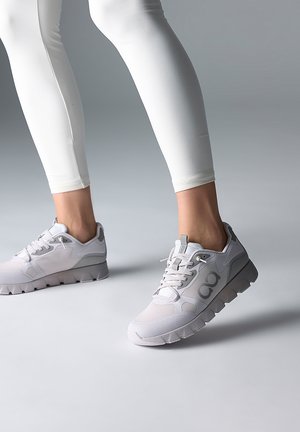 Beine in weißen Leggings, die weiße und graue Sportschuhe tragen und auf einer glatten, neutral-grauen Oberfläche stehen.