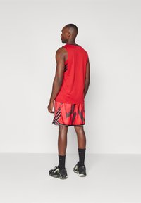 Rotes ärmelloses Sportoberteil mit schwarzen Akzenten, kombiniert mit roten gemusterten Shorts, die diagonale schwarze Formen aufweisen. Schwarze Sportschuhe und Socken.
