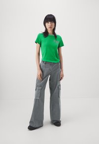 T-shirt in cotone verde con dettagli bianchi e logo, abbinato a pantaloni cargo grigi a gamba ampia con tasche laterali e una consistenza morbida.
