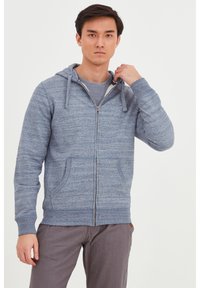 Haut zippé gris en tissu texturé, avec une capuche à cordon, des poches avant et des poignets côtelés, porté sur une chemise grise.