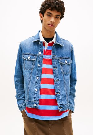 Jeune homme aux cheveux bouclés portant une veste en jean bleue par-dessus un maillot de rugby rayé rouge et bleu clair et un pantalon marron.