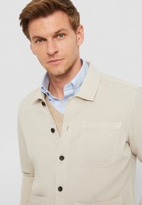 Giacca camicia in cotone beige con colletto, chiusura a bottone, tasca sul petto singola e bottoni scuri, indossata sopra una camicia a righe azzurro chiaro.