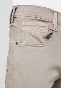 Jeans de mezclilla beige con una textura suave, que cuentan con un bolsillo trasero con un diseño en forma de V y un cierre de botón metálico.
