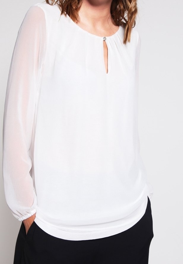 Femme portant un chemisier blanc à manches longues avec une petite ouverture en forme de goutte à l'avant et un pantalon noir, mains dans les poches, sur un fond uni.