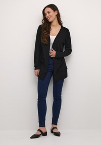 Cardigan nero con maniche lunghe, apertura frontale e due tasche frontali, abbinato a una maglietta bianca e jeans blu. Il tessuto è morbido con una texture liscia.
