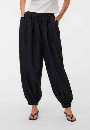 Femme portant un pantalon noir ample à taille élastique avec des bas froncés et des sandales noires à talons, debout les mains dans les poches.