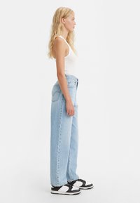 Vaaleahiuksinen nainen, joka pukeutuu valkoiseen hihattomaan paitaan, vaaleansineisiin korkeavyötäröisiin farkkuihin ja musta-valkoisiin tennareihin, seisoo profiilissa yksinkertaista valkoista taustaa vasten.