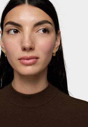 Donna con capelli lunghi e scuri che indossa piccoli orecchini a cerchio dorati e un top marrone a collo alto, che guarda leggermente verso destra contro uno sfondo uniforme.