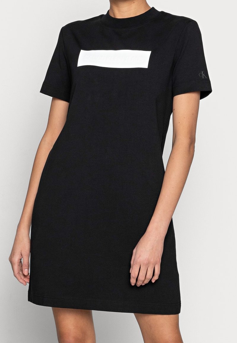Femme portant une robe t-shirt courte noire Calvin Klein avec un logo rectangulaire blanc sur la poitrine et les initiales CK sur la manche.