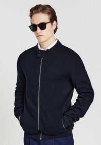 Maglione zip-up blu scuro con colletto alto, scollatura rotonda e polsini a costine. Materiale in maglia leggero, con chiusura a due bottoni.