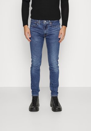 Man draagt slim-fit blauwe jeans, een zwart langemouwen top en zwarte veterschoenen, staand tegen een effen witte achtergrond.