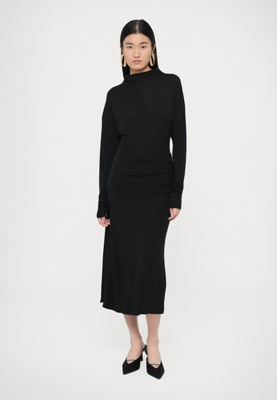 Fekete, hosszú ujjú ruha, testhezálló derékrésszel, redőzött dizájnnal és magas nyakkivágással, sima, rugalmas anyagból készült. Magassarkú fekete cipők.