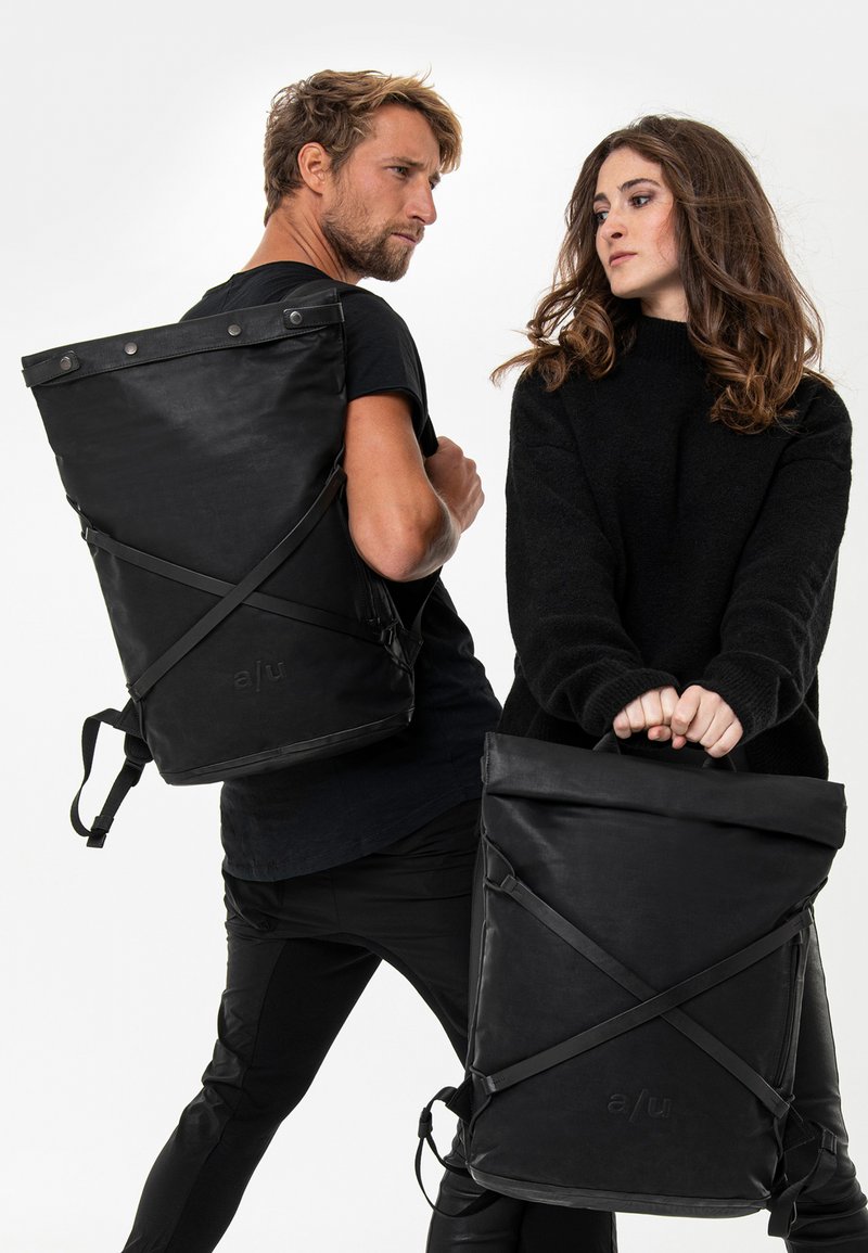 Zwarte roll-top rugzakken met verstelbare straps, voorzien van een matte afwerking en zichtbaar logo. Eén wordt gedragen, de andere wordt vastgehouden door een model.