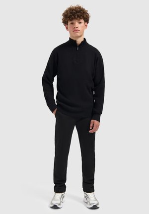 Schwarzer Pullover mit Reißverschluss aus strukturiertem Stoff, kombiniert mit schwarzen Hosen und silbernen Sneakers. Das Modell steht vor einem neutralen Hintergrund.