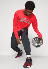 Ung mand i rød Chicago Bulls sweatshirt og sorte bukser, der dribler en sort-hvid basketball mod en ensfarvet baggrund.