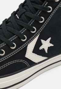 Zapatillas altas de tela negra con logo de estrella de cuero blanco, costuras en contraste, ojales de metal y suela de goma con franjas blancas y negras.