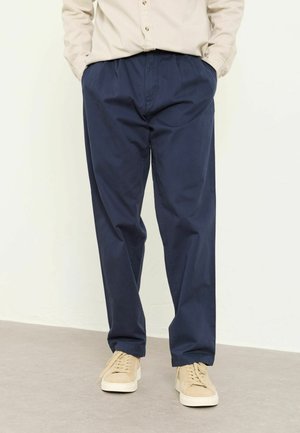 Mann trägt marineblaue Freizeithose, hat die Hände in den Taschen und beige Sneaker auf grauem Boden vor weißer Wand.