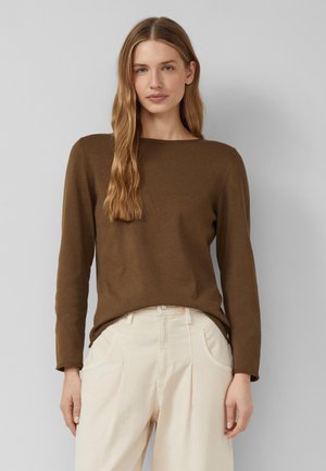 Młoda kobieta o długich blond włosach, ubrana w brązową bluzkę z długim rękawem i kremowe, wysokie, szerokie spodnie, stoi na jednolitym tle.