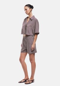 Mujer con camiseta corta de rayas verticales en color marrón y blanco y pantalones cortos a juego, de pie de perfil con sandalias planas negras.