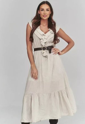 Kobieta o długich brązowych włosach, ubrana w beżową, marszczoną sukienkę midi z czarnym paskiem, stojąca z jedną ręką na biodrze na jednolitym tle.