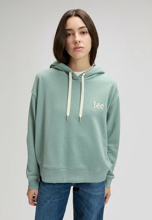 Nuori nainen, jolla on tummat hiukset ja joka pukeutunut vaaleanvihreään Lee-huppariin ja sinisiin farkkuihin, seisoo yksinkertaista vaaleaa taustaa vasten.